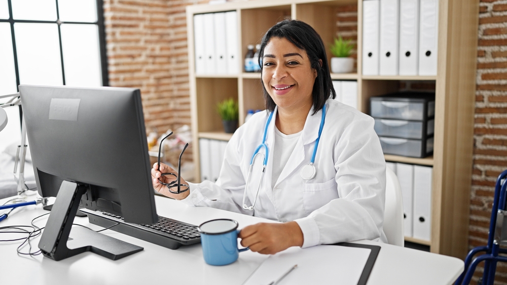 A smiling doctor sits at a desk with a computer in a clinic office setting