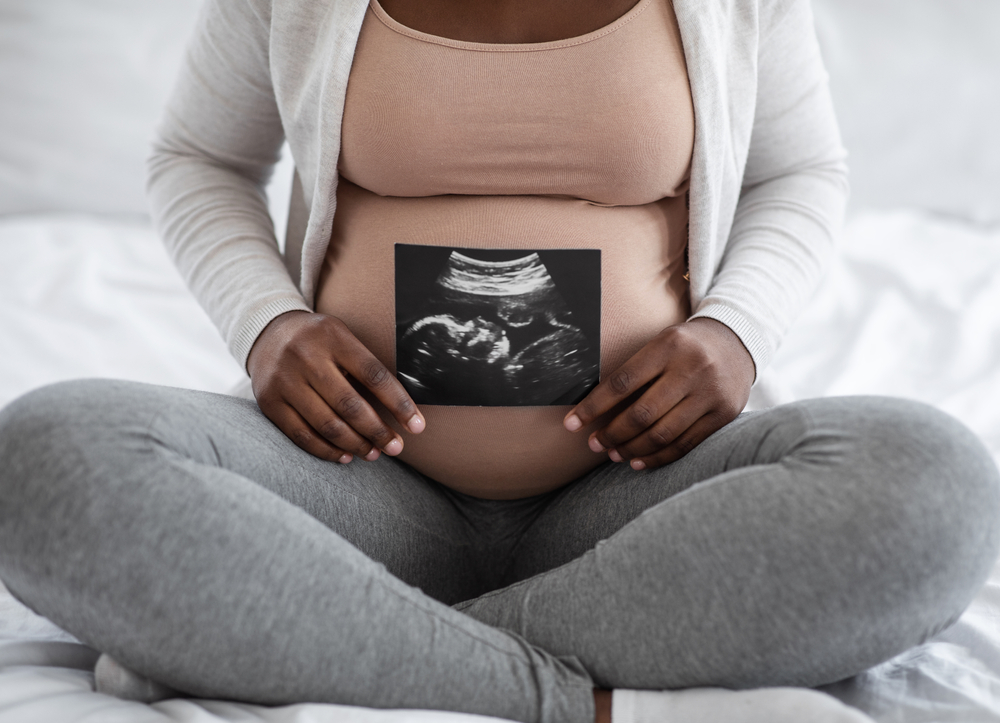 Unrecognizable black pregnant lady demonstrating her baby sonography photo while sitting on bed at home